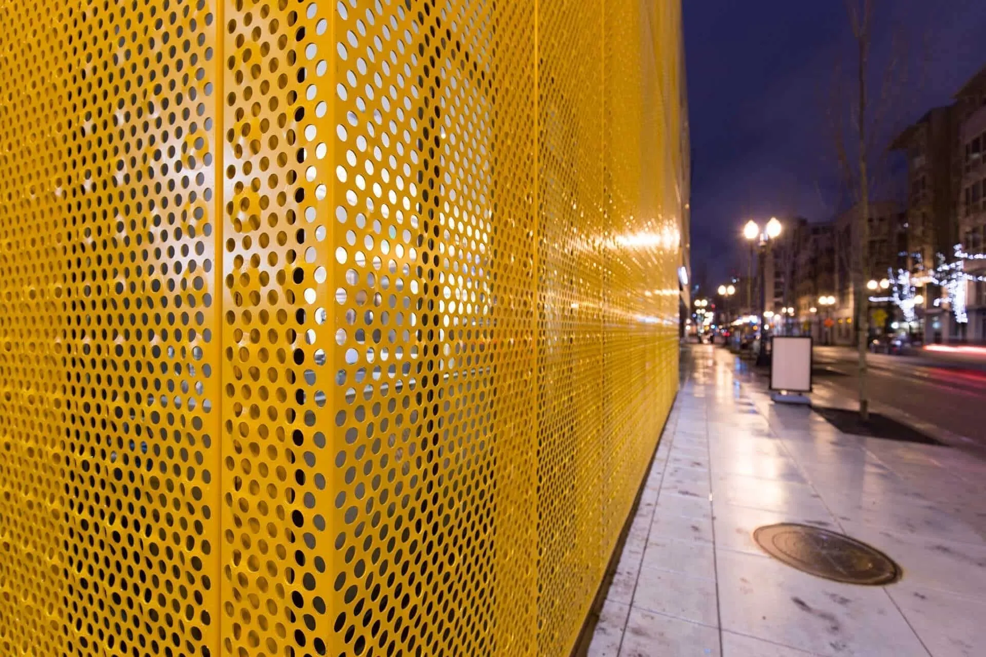 Close on the perforated screen’s corner, the wet sidewalk and streetlights beyond