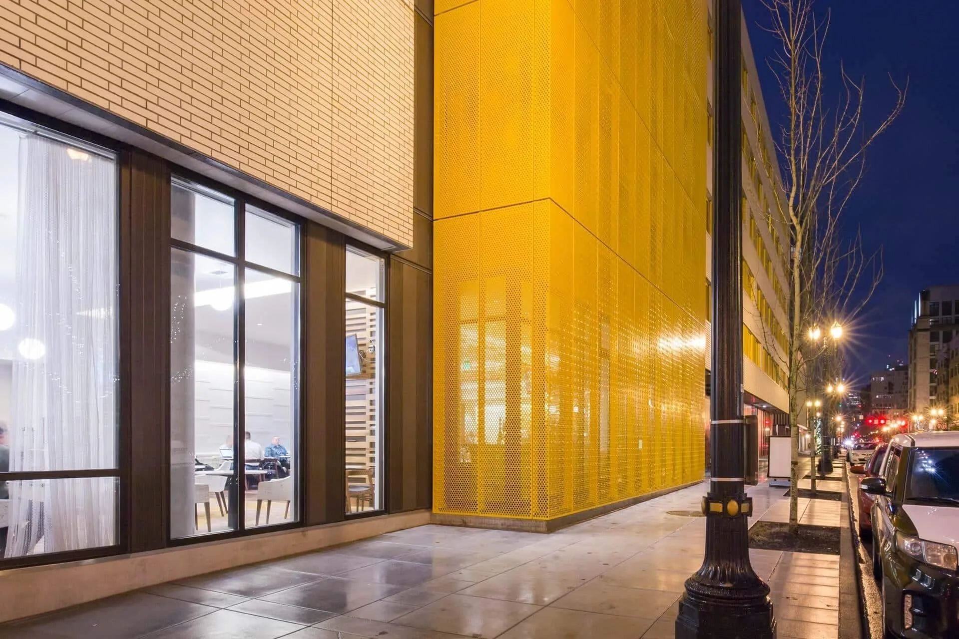 Along the sidewalk at dusk, lobby windows giving way to the perforated yellow screen