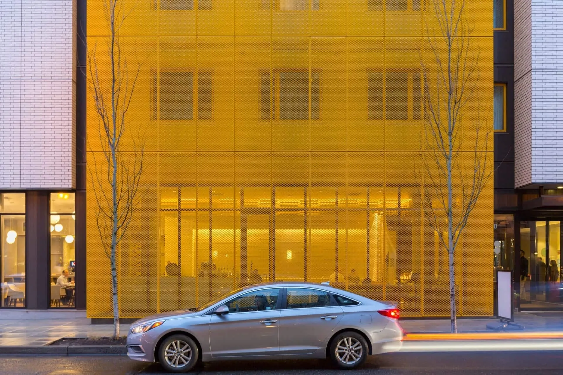 The yellow screen wall over the lit ground floor, a silver car passing on the street