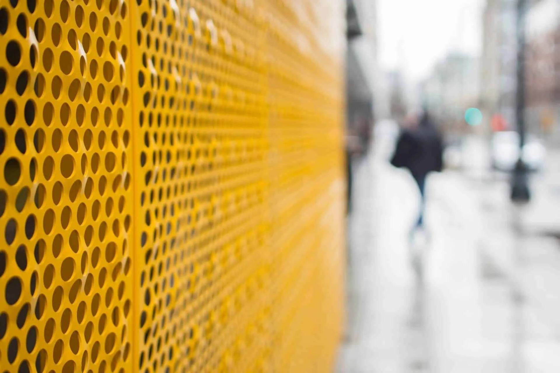 Round perforations in the yellow screen, a pedestrian blurred on the sidewalk behind