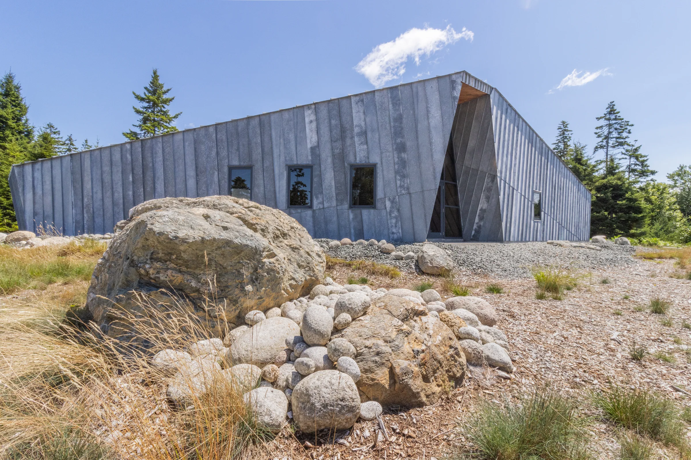 A long zinc wall rising behind boulders and beach stones