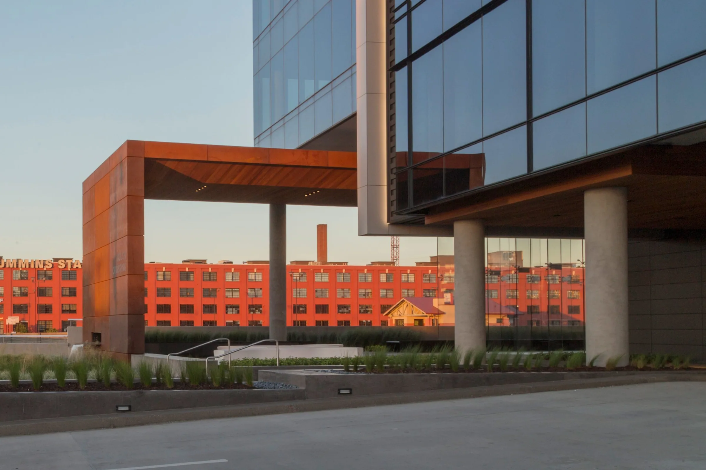 A rust-colored steel canopy beside the glass tower at sunset
